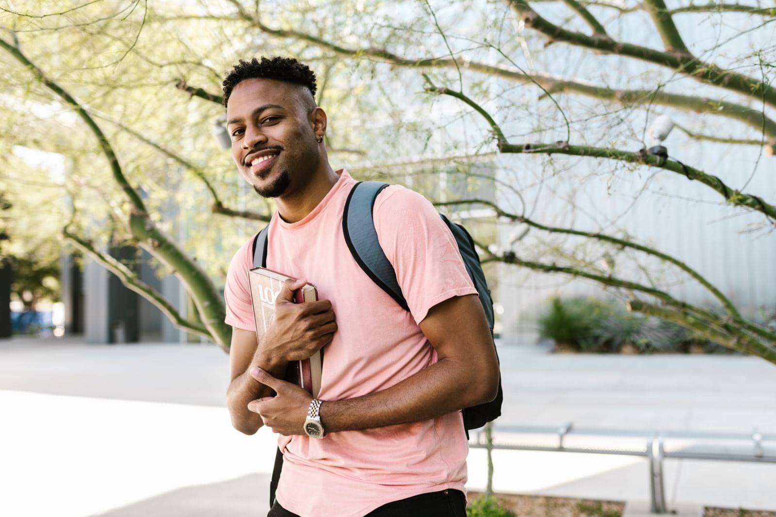 A student with a backpack holding a book, smiling, standing outdoors among trees.