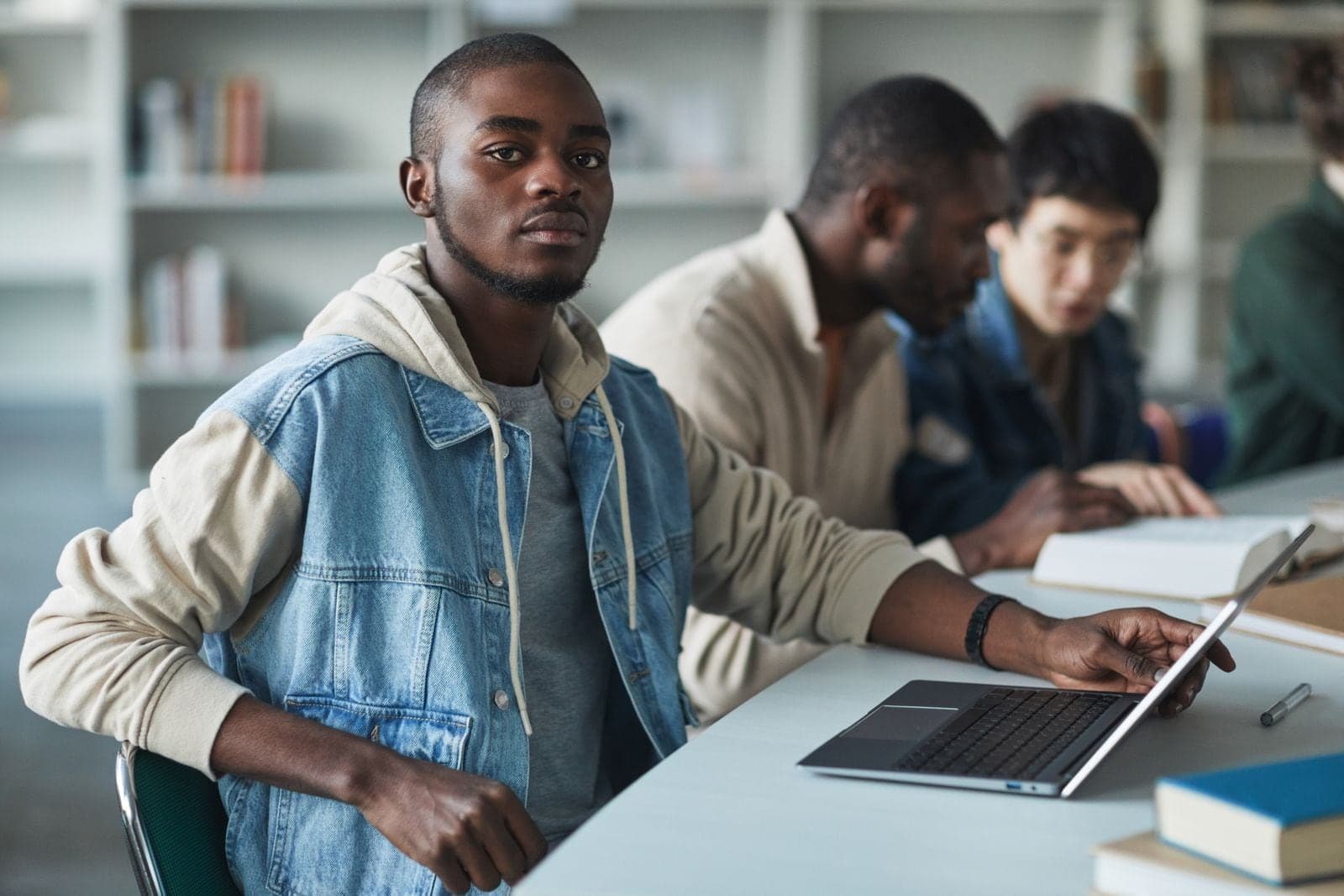 A student sitting at a long table in a library, with others working at laptops and books beside them.