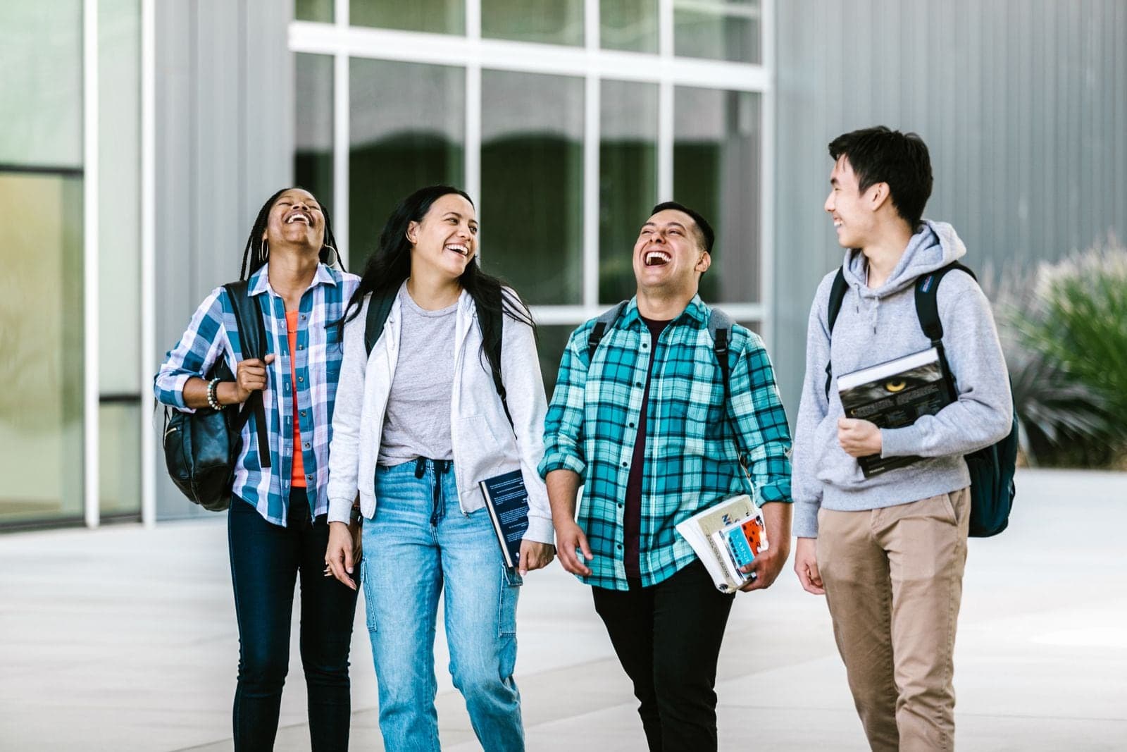 Four students walking together outside a building, carrying books and bags, laughing.