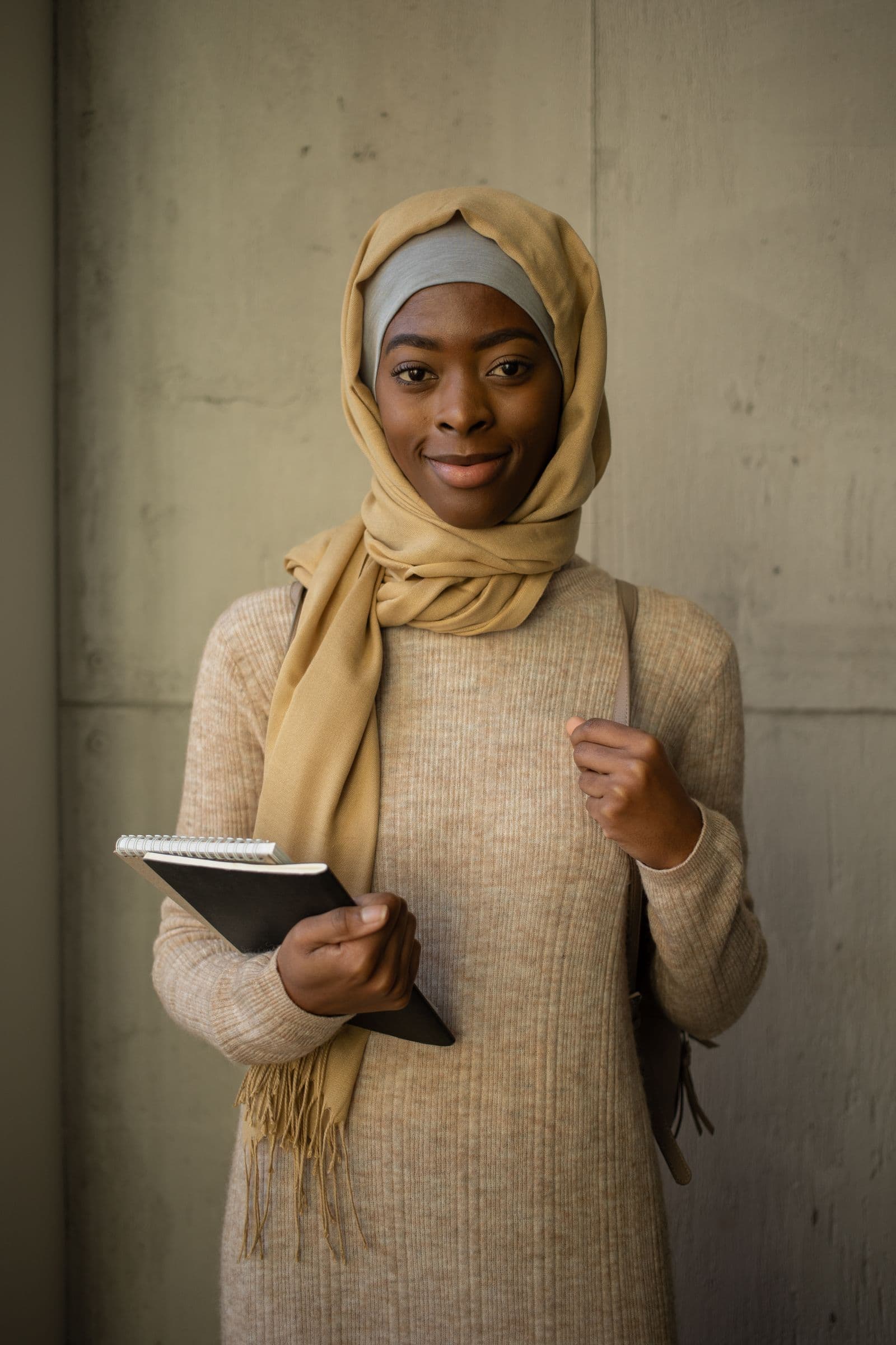 A student holding a notebook with a backpack over her shoulder, standing against a concrete wall.