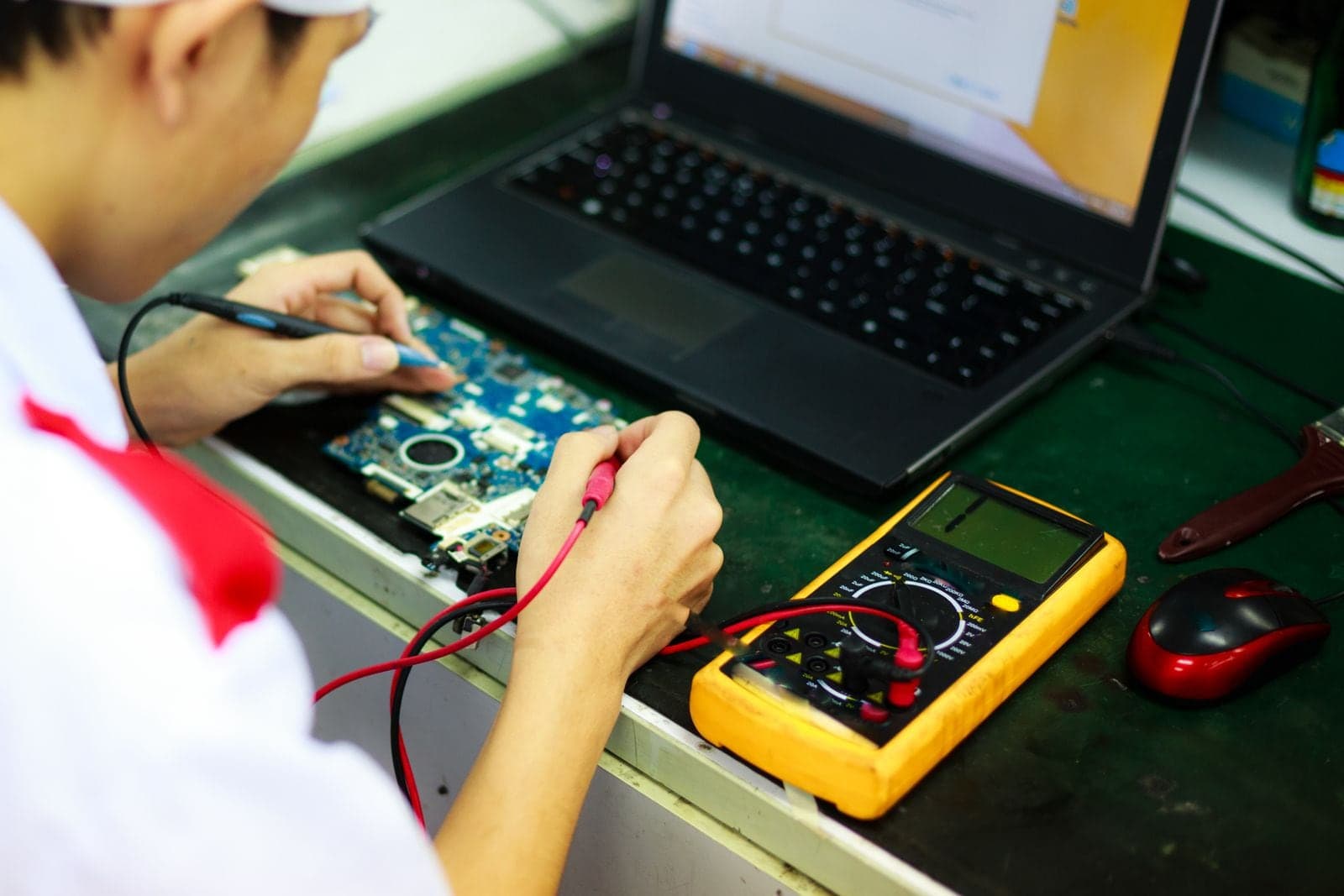 A technician testing a laptop circuit board with a multimeter at a workbench.